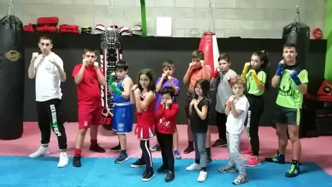 Grupo de niños y niñas del club posando en guardia junto a su entrenador, delante de los sacos del gimnasio